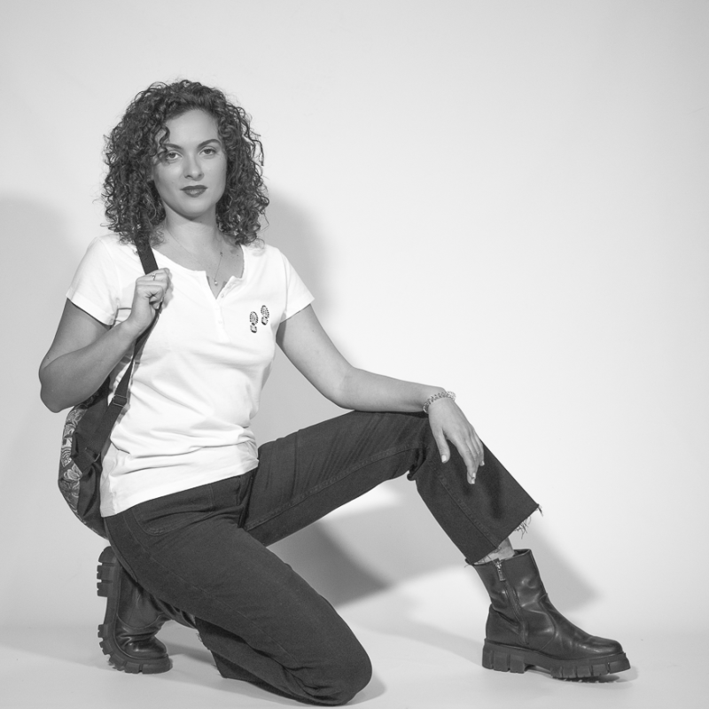 Modelo en estudio con camiseta Ripariam blanca de cuello henley y estampado de huellas en el pecho, combinada con pantalones negros y botas de suela gruesa, posando con mochila sobre fondo neutro en blanco y negro.