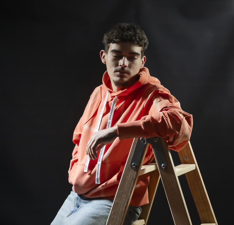 Modelo en estudio luciendo sudadera Ripariam estilo tie-dye en tono terracota con cordones personalizados, posando apoyado en una escalera de madera sobre fondo oscuro.