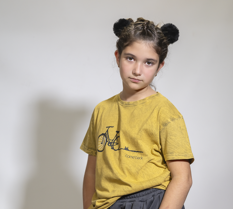 Niña posando en estudio con camiseta amarilla estampada con una bicicleta y el texto “come back”, sobre fondo neutro.