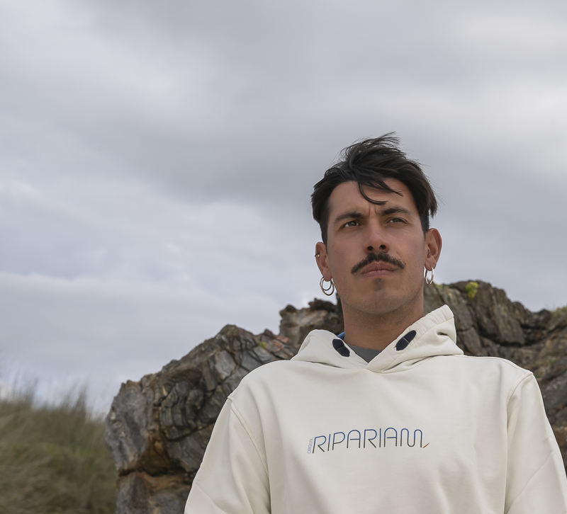 Modelo al aire libre con sudadera Ripariam blanca y capucha, posando frente a formaciones rocosas bajo un cielo nublado.