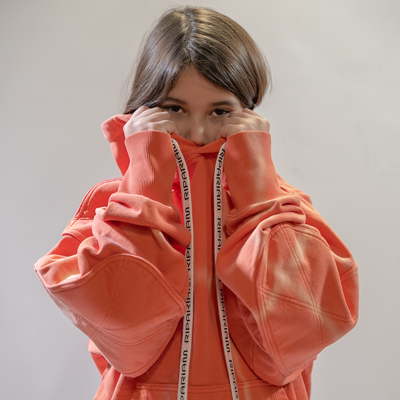 Niña con sudadera naranja Ripariam, posando con capucha ajustada y detalles de cuerda de logo, ropa urbana y cómoda