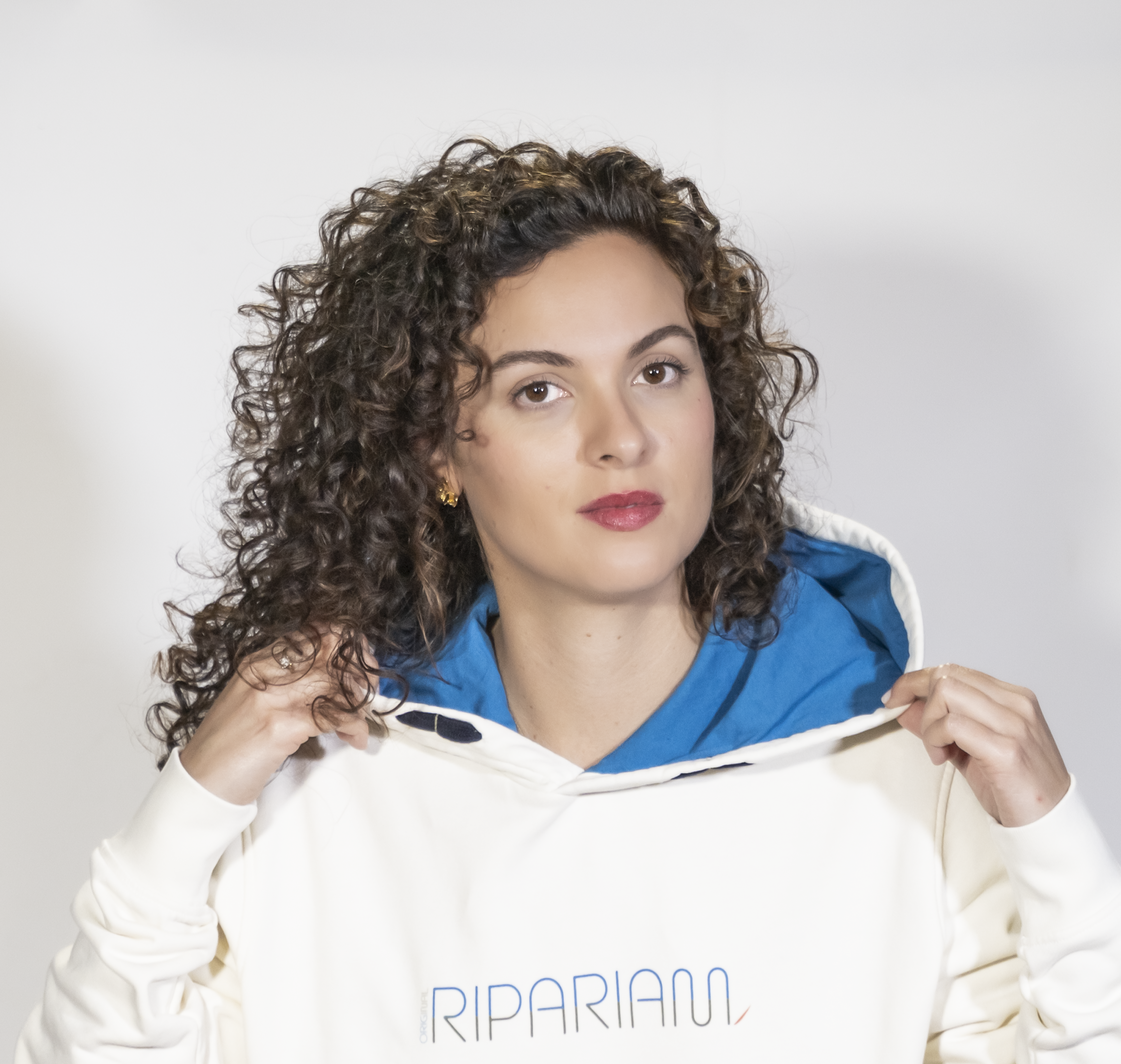 Mujer con sudadera blanca Ripariam, posando con capucha azul, detalles de logo moderno en el pecho, imagen de moda urbana y sostenible