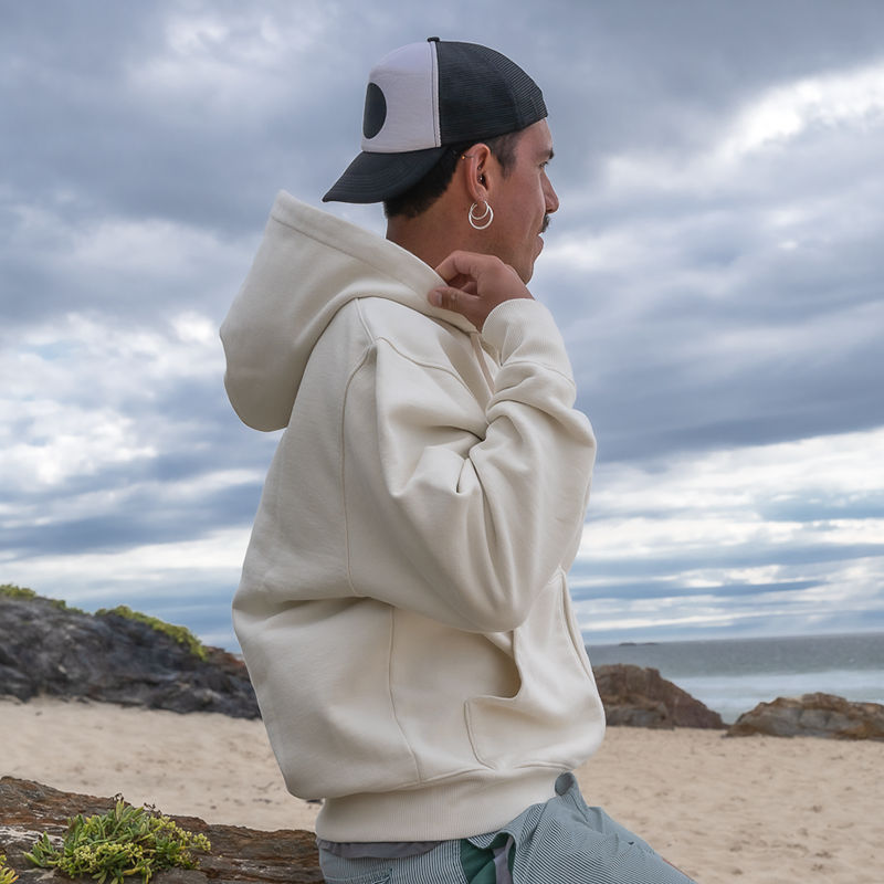 Hombre con sudadera blanca y gorra negra, posando en la playa frente al mar, imagen en ambiente relajado y estilo urbano